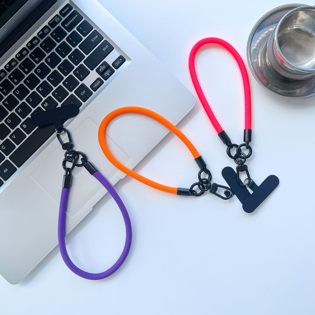 Três cordões de pulso para capa de celular nas cores roxo, laranja e rosa neon, dispostos sobre um notebook e ao lado de uma xícara de metal.